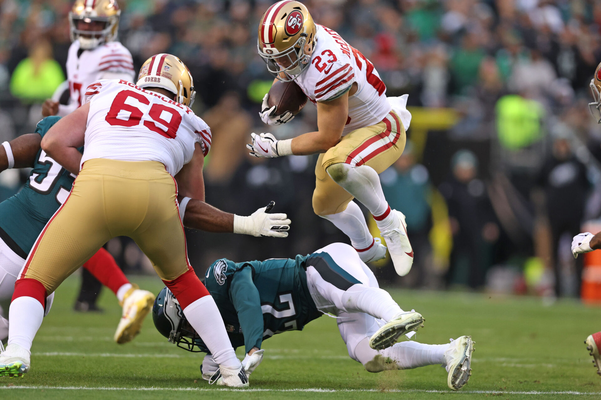 Christian McCaffrey, in a white jersey, gold pants and a gold helmet (all with red accents) hurdles an Eagles player (in a green jersey)
