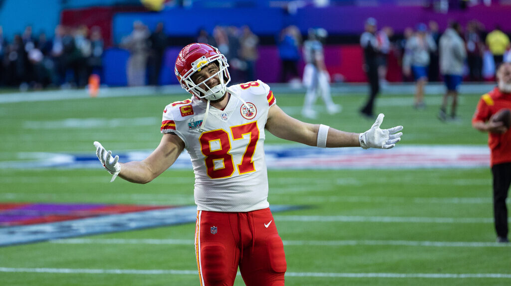 Kansas City tight end Travis Kelce poses and smiles at the camera before the start of a game. He's standing on the field wearing a white jersey, red helmet and red pants.
