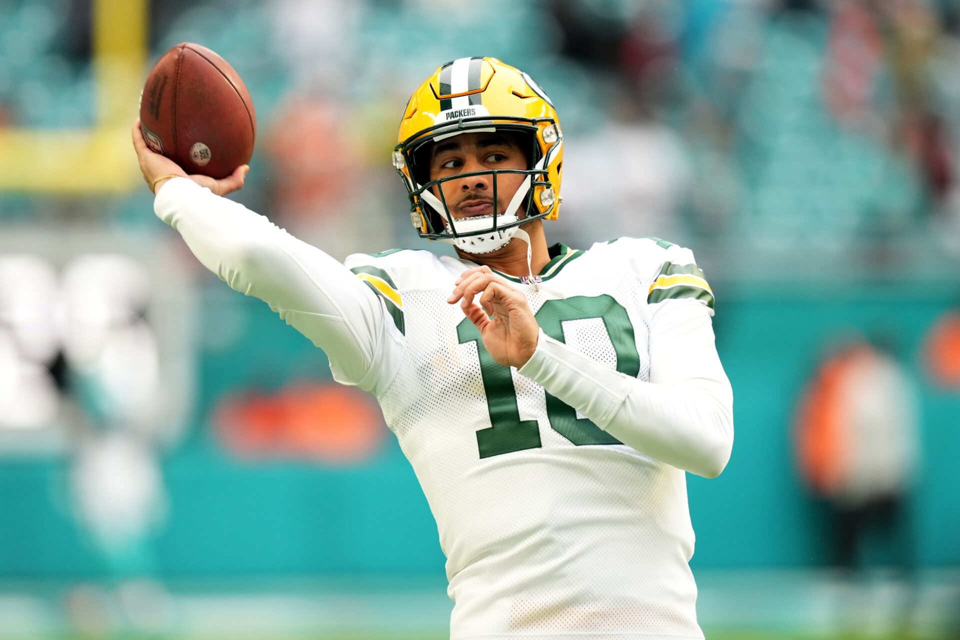 Green Bay Packers QB Jordan Love throwing a pass in focus in front of a blurry background. He's wearing a white jersey with a white long-sleeve underneath and a yellow helmet.