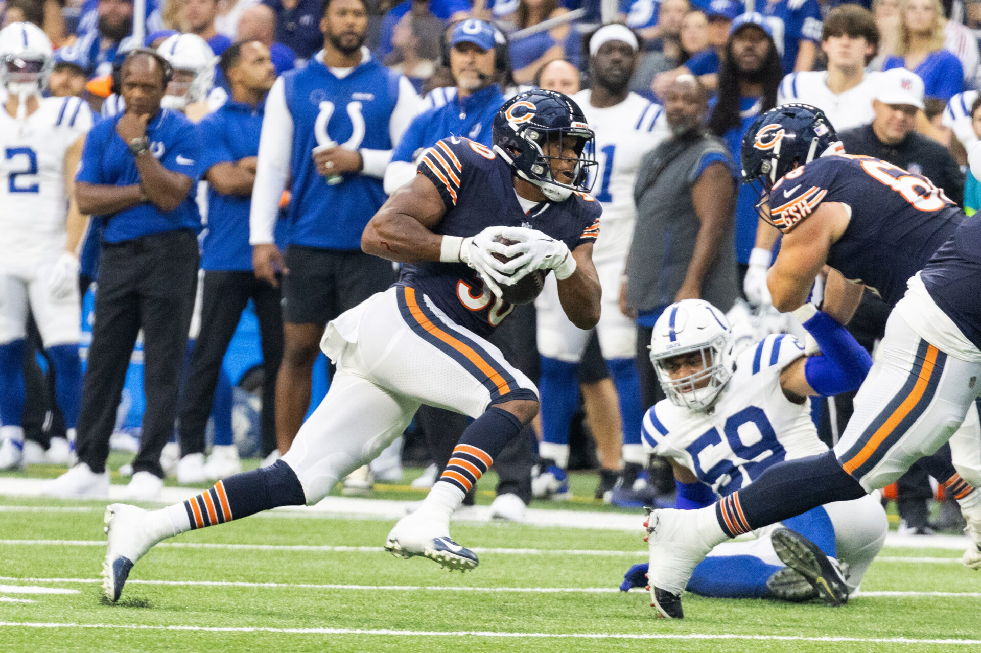 Roschon Johnson, in a dark blue jersey and helmet and white pants, runs past a Chicago teammate and a Colts defender on the ground