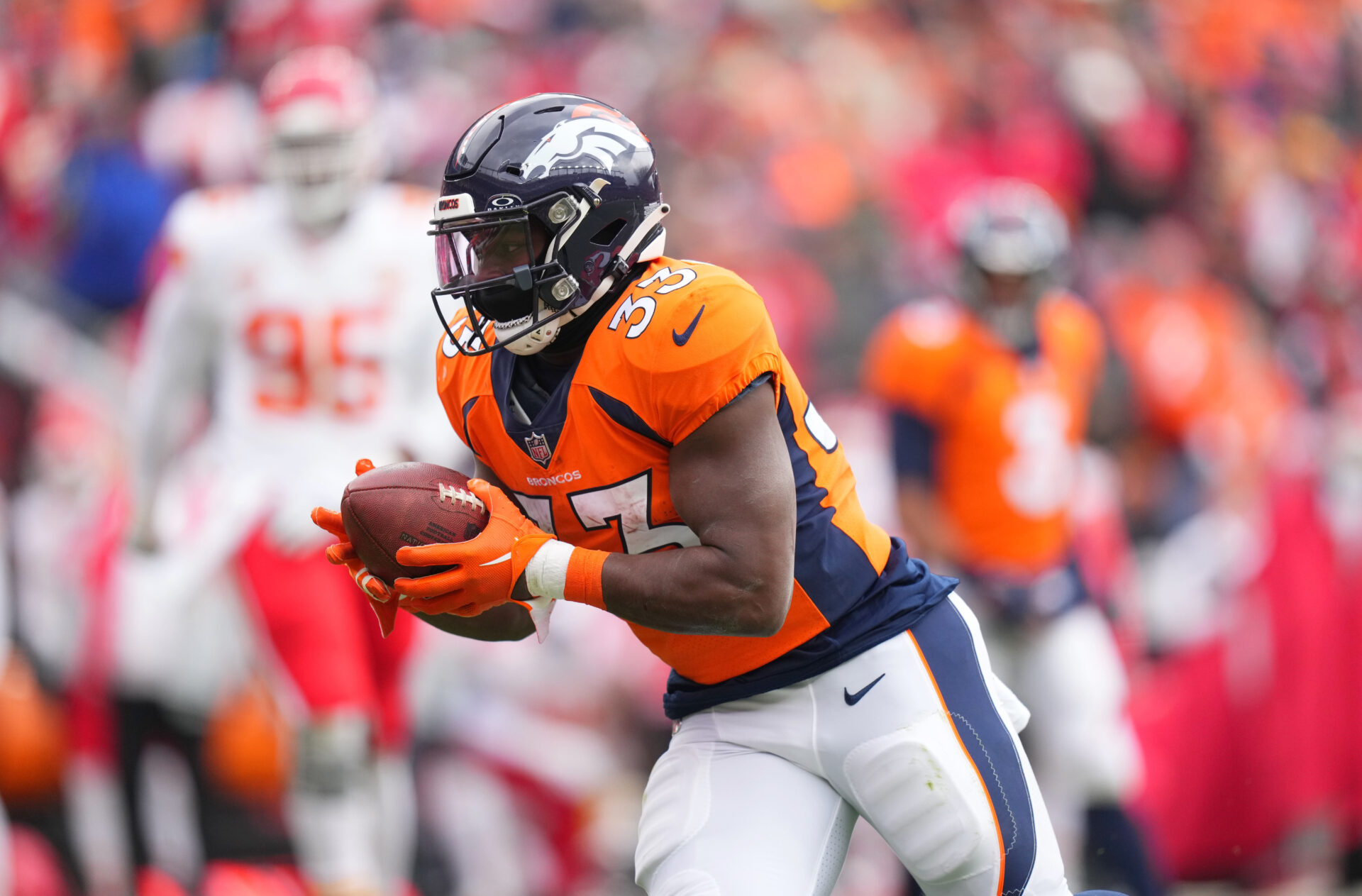 An above-the-knees image of Javonte Williams, in an orange Denver Broncos jersey and white pants, running the football with the rest of the image's background blurry