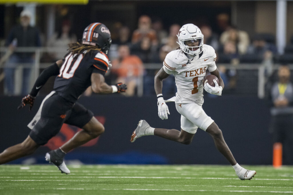 Xavier Worthy, in an all-white uniform, looks to evade an Oklahoma State defender (in black)