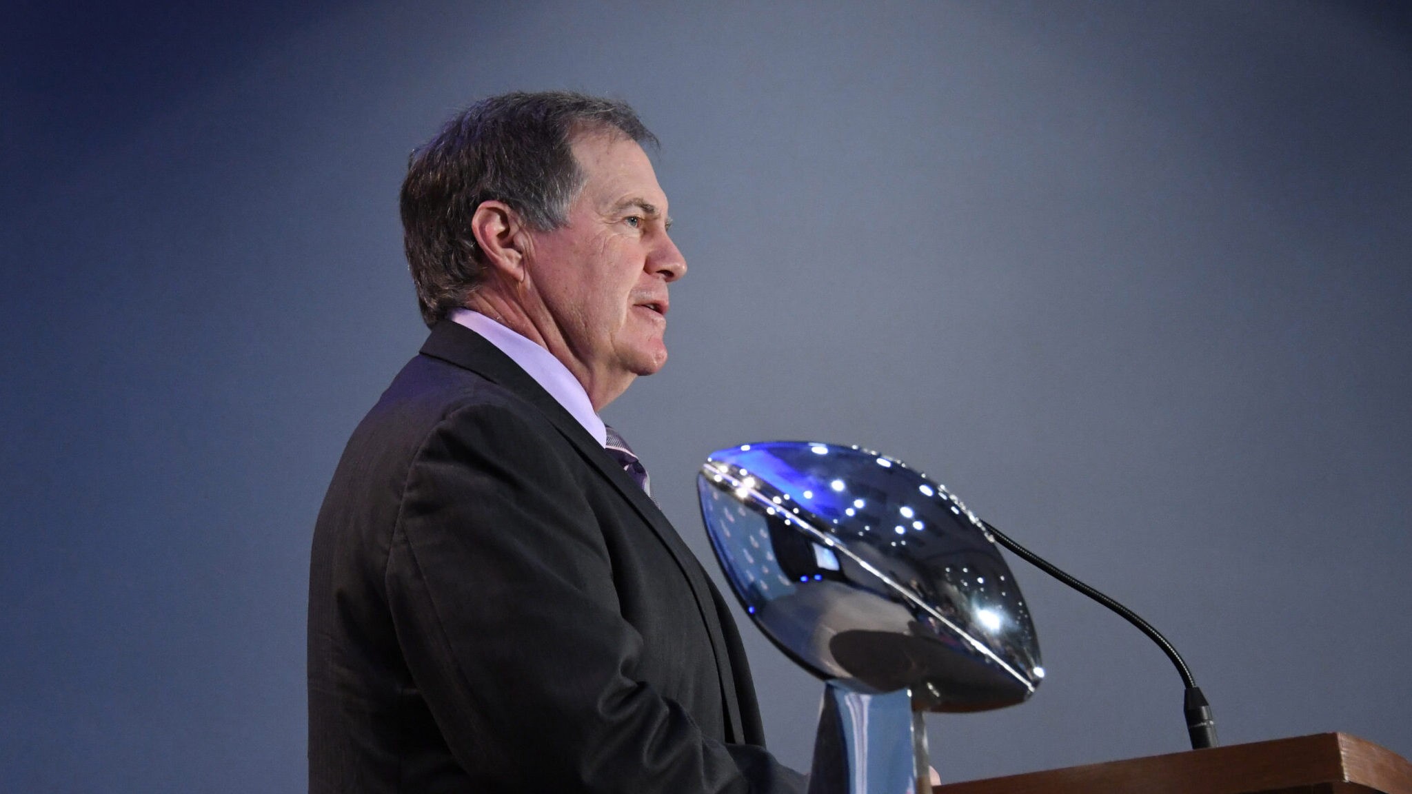 Bill Belichick stands at a podium with the Super Bowl trophy in the shot.