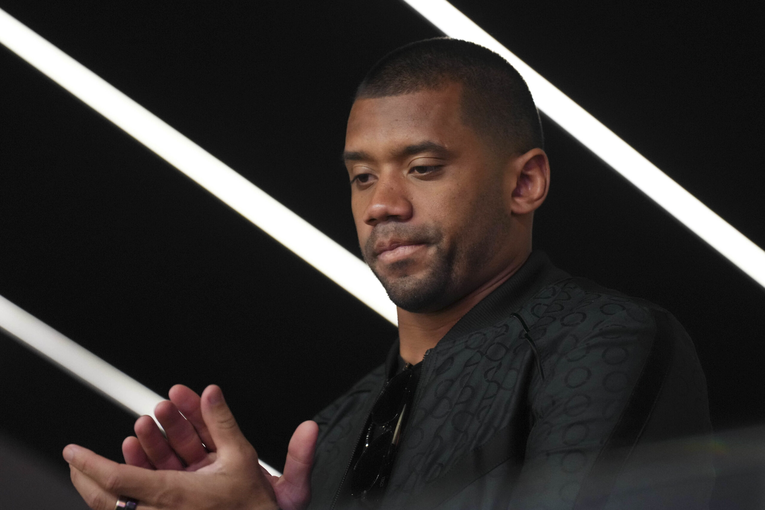 Russell Wilson looks downcast in front of a black-and-white-striped background in a black jacket and shirt