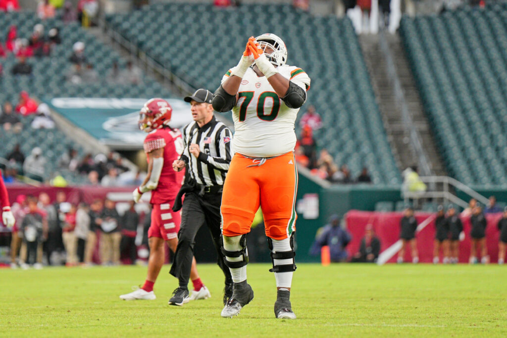 Javion Cohen, in a white jersey and helmet and orange pants, raises his hands as he stands behind the play