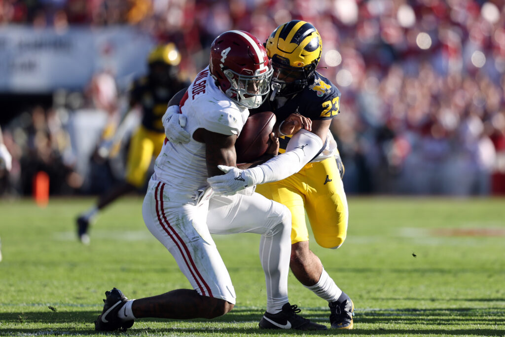 In a yellow helmet, blue jersey, and yellow pants, Michigan linebacker Michael Barrett sacks Alabama's quarterback (dressed in white with a red helmet)