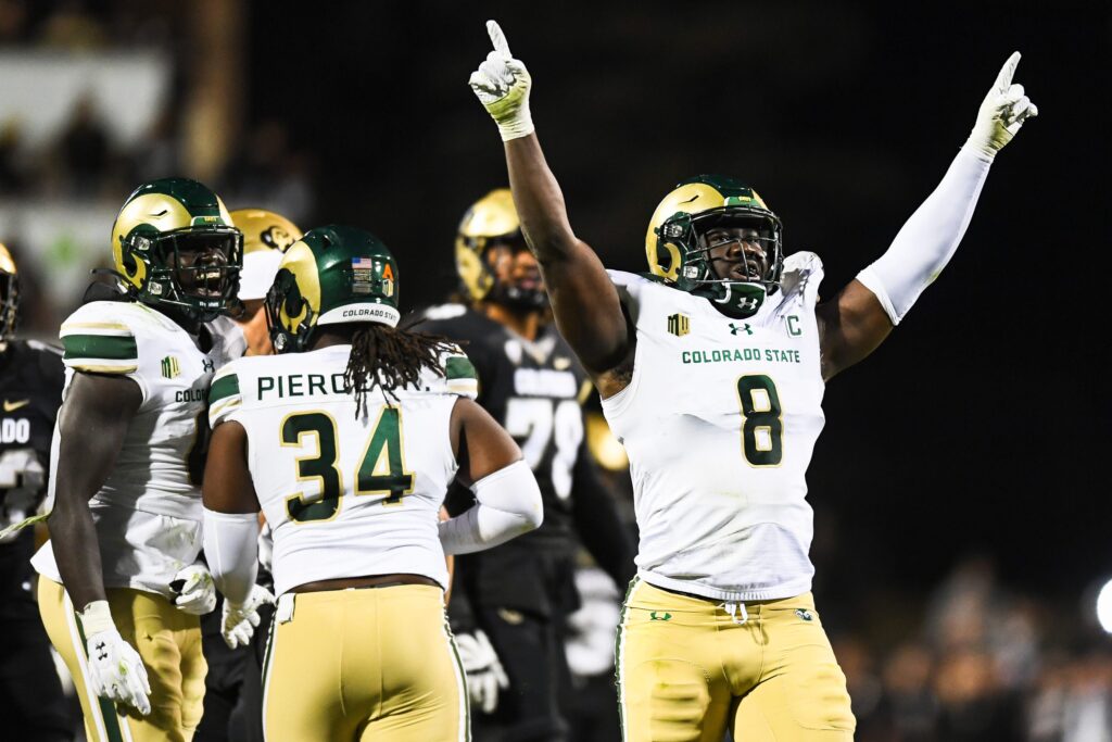 Colorado State defensive end Mohamed Kamara (in a white jersey, light yellow pants, and a green-and-gold helmet) raises his arms up in front of a completed play
