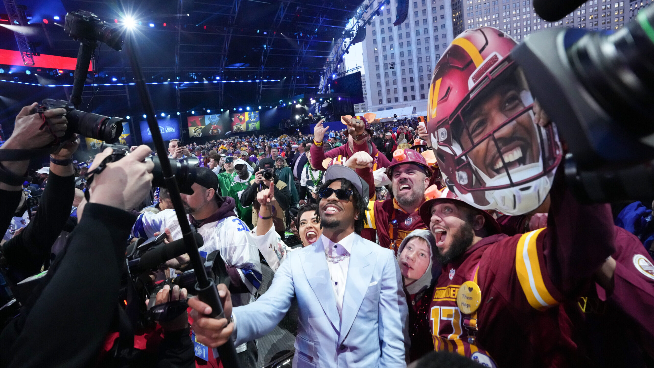 Washington Commanders quarterback Jayden Daniels (in a pale blue suit) stands among fans and cameras