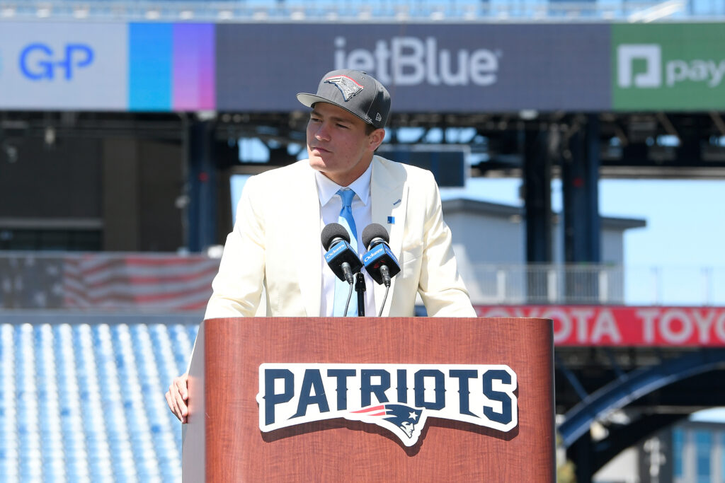Upper-body image of New England Patriots quarterback Drake Maye (in a cream suit jacket, white shirt, cerulean tie and gray Patriots hat) stands behind a podium