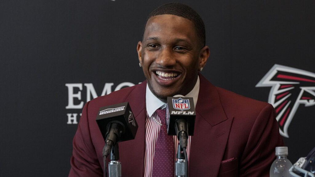 Upper-body image of Atlanta Falcons quarterback Michael Penix Jr. (in a maroon suit and tie and light red shirt) seated at a table with microphones in front of a black background with the Falcons' logo