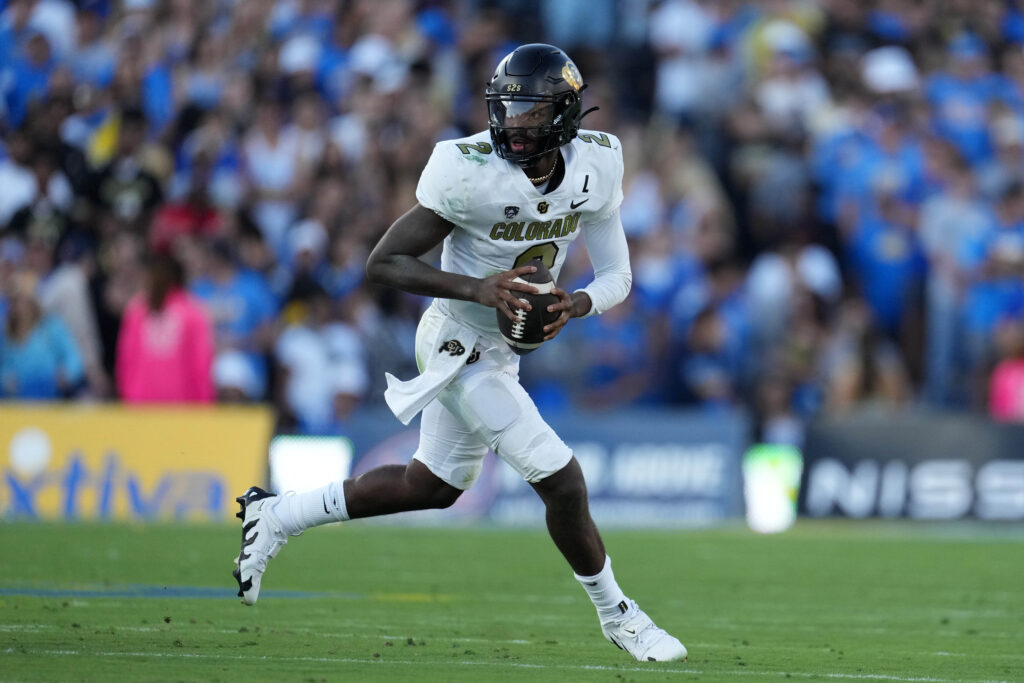 Colorado quarterback Shedeur Sanders drops back to throw. He's in a white uniform and black helmet and is the only player in frame with the crowd blurry behind him