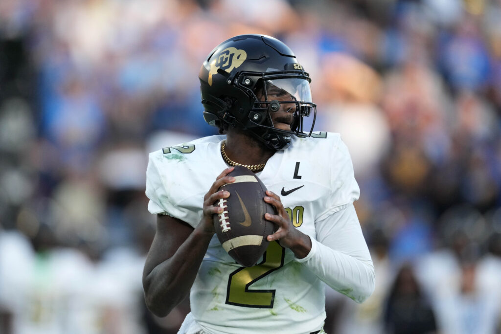 Upper body image of Colorado quarterback Shedeur Sanders with the ball in his hands, looking downfield to throw. He's in a white jersey and black helmet